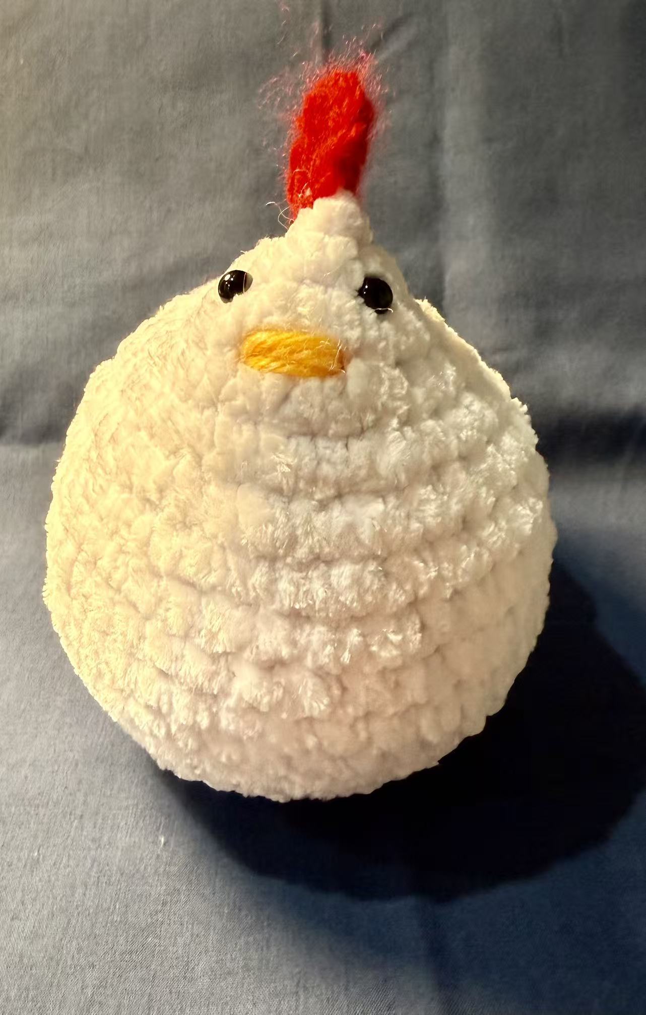 Front view of a white crochet chicken with a black eye, a yellow stitched beak, and a red comb.